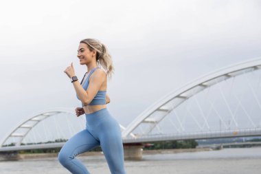 Spor giyimli genç bir kadın iskelede koşuyor ve gülümsüyor. Sağlıklı yaşam tarzı, spor yaşam tarzı. Arka planda bir köprü var.