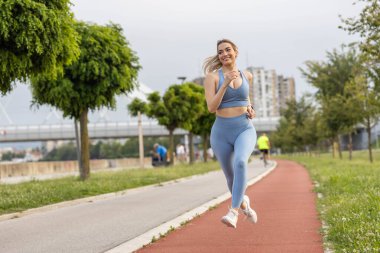  Şehirdeki yarış pistinde koşan spor giysili neşeli genç bir kadın.