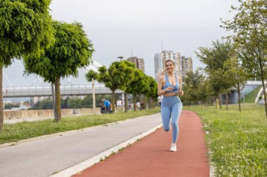  Şehirdeki yarış pistinde koşan spor giysili neşeli genç bir kadın.