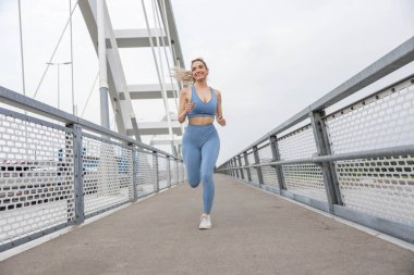 Köprüde koşan güzel, sportif genç bir kadın. Güçlü vücut ve bacaklara sahip kadın koşucu köprüden ve eğitimden geçiyor. Spor ve eğlence..