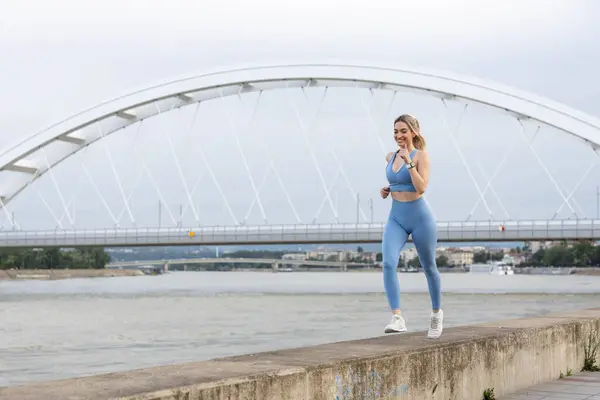 Spor giyimli genç bir kadın iskelede koşuyor ve gülümsüyor. Sağlıklı yaşam tarzı, spor yaşam tarzı. Arka planda bir köprü var.