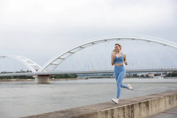 Spor giyimli genç bir kadın iskelede koşuyor ve gülümsüyor. Sağlıklı yaşam tarzı, spor yaşam tarzı. Arka planda bir köprü var.