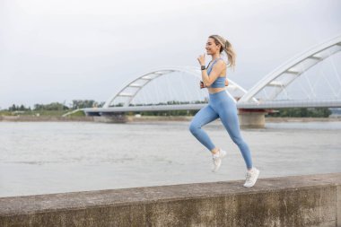  Spor giyimli genç bir kadın iskelede koşuyor ve gülümsüyor. Sağlıklı yaşam tarzı, spor yaşam tarzı. Arka planda bir köprü var.