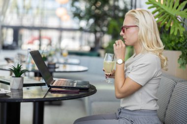 Sarışın bir kadın, yeşil ve doğal ışıkla dolu modern ve rahatlatıcı bir atmosferi olan bir kafede laptopuyla çalışırken içkisinden bir yudum alır..