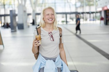 Gezgin, coşkulu bir heyecanla, hareketli bir tren istasyonunun ortasında, doğal ışıkla parlayan, beklenen bir maceraya atılmaya hazır, buzlu bir kahve tutuyor..