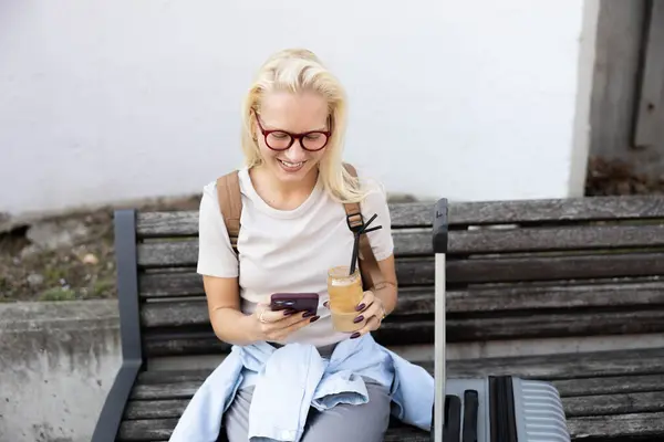 Sarışın bir kadın gezgin havaalanındaki bir bankta oturuyor, akıllı telefonunu kullanıyor ve yanında bir bavulla, seyahatlerine hazırlanıyor..