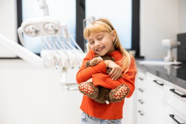 Diş kliniğinde genç bir kız oyuncak ayısına sarılıyor klinik bir ortamda rahatlık ve sakinlik gösteriyor ve pediatrik hasta bakımını ön plana çıkarıyor..