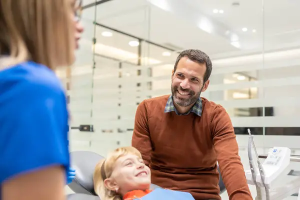 Bir baba ve çocuk dişçi muayenehanesindedirler, çocuğun kliniğe diş muayenesi sırasında ebeveynlik desteğinin ve güven inşa etmenin önemini vurgularlar..
