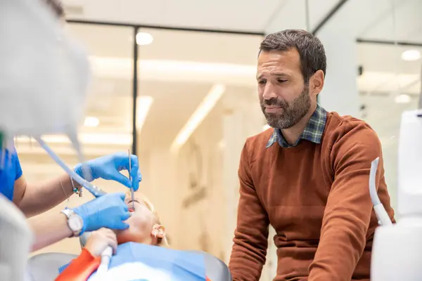 Çocuk diş sağlığı kontrolünden geçerken, modern bir diş sağlığı kliniğine yapılan ziyaretlerde aile desteğini ve endişelerini vurgulayan bir baba endişeli görünür..