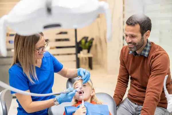 Baba, çocuğun diş tedavisini izlerken gülümsüyor, modern bir klinik diş ortamında ebeveyn güvencesi ve konfor anı yakalıyor..