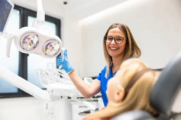 Gözlük ve eldiven takan bir dişçi, genç hastasıyla modern ve parlak bir diş kliniğinde etkileşime girer..