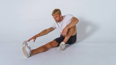 Sıradan tişörtlü, siyah pantolonlu, beyaz arka planda esneme hareketleri yapan sarışın genç yakışıklı bir adam. Spor konsepti