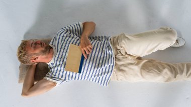 Yakışıklı sarışın bir adam, gündelik giysiler içinde elinde kitapla yerde yatıyor. Rahatla konsept