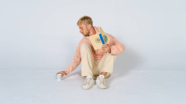 Beyaz arka planda sıradan giyinmiş yakışıklı sarışın bir adam yerde oturmuş kitap okuyor ve içki molası veriyor. Rahatla konsept