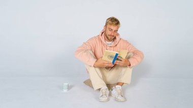 Beyaz arka planda rahat giyinen yakışıklı sarışın bir adam bir yastığın üzerinde oturup kitap okuyor ve içki içiyor. Entelektüel kavram