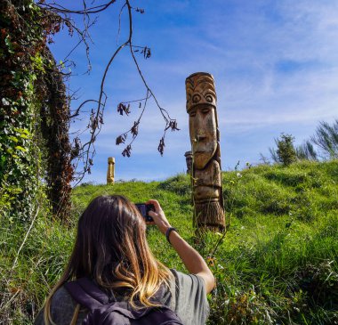 Uzun saçlı genç bir kadın yeşil çimenlerde çömelmiş, ahşap bir moai 'nin fotoğrafını çekiyor. Heykel yüksek bir konumda..