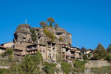 Rupit kasabasının en yüksek kesimindeki evler ve taş binalar. Evlerin üzerinde, sarp bir kayanın tepesinde birkaç ağaç ve bağımsız bir Katalan bayrağı var.