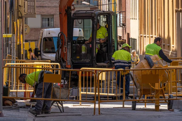 Stock image Workers working on public works in a street in Granollers. Protection fences and backhoe. Passersby walking. Concrete mixer.;