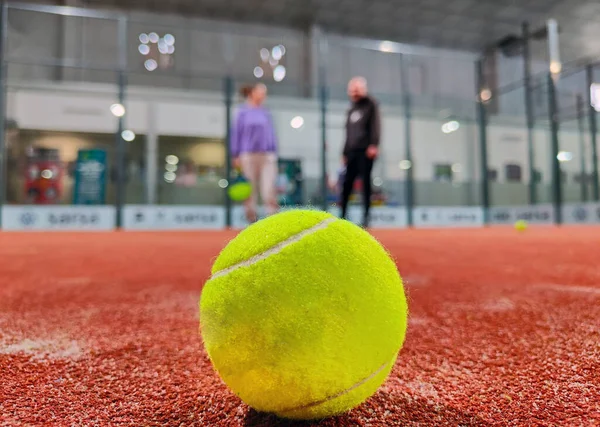 Detail shot of a paddle ball on the floor of the indoor paddle tennis court and three player friends in the background of the court