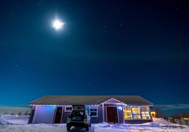 Rural house beautifully illuminated with colorful Nadal lights and a woman protected from the cold inside an open trunk under a starry sky with a green aurora borealis and a completely snowy environment with trees