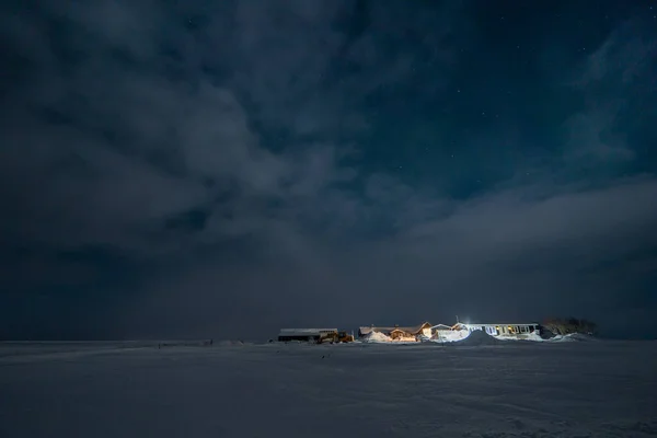 Isolated illuminated farmhouse with completely snowy surroundings under a sky with clouds and clear at night with stars and a faint green light of aurora borealis.