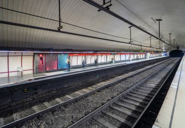 Train tracks and rails of the Barcelona metro or subway station and passengers waiting seated or walking with a train appearing from the beginning of the tunnel.