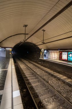 Train tracks and rails of the Barcelona metro or subway station and passengers waiting seated or walking with a train appearing from the beginning of the tunnel.