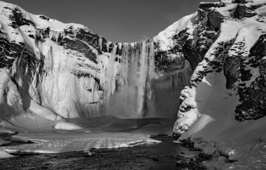 Giant skgafoss waterfall completely covered with snow and the river passes next to it. The dawn sun illuminates the water that falls from the waterfall in black and white.