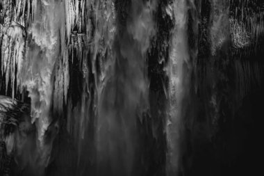 Detail of stalagmites and water falling from the skgafoss waterfall in Iceland illuminated by late afternoon sunlight creating a painterly atmosphere as if it were an abstract black and white artwork