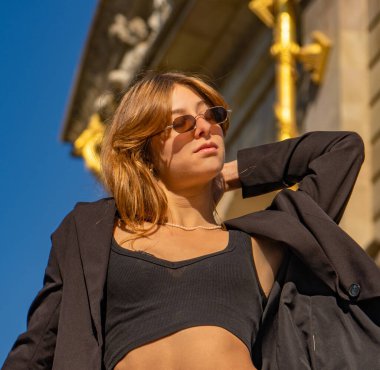 Uzun sarı saçlı, güneş gözlüklü, siyah bluzlu güzel bir manken siyah ceketle güneşleniyor ve saçlarına dokunuyor.