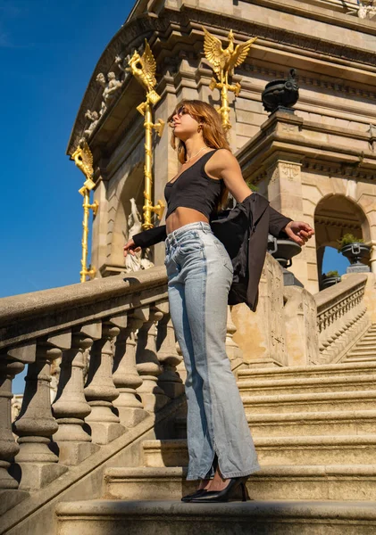 Beautiful young fashion model long blond hair in blue jeans, sunglasses, black top and blue jeans standing on a monument staircase with open arms and black jacket at elbows with open arms sunbathing