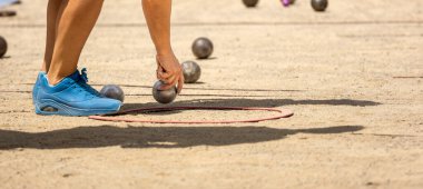 Spor ayakkabılı genç bir kadının eli, kumlu bir Petanque kortunda, güneşli bir günde Petanque oyunu oynamak için metal petanque topunu yakalıyor.