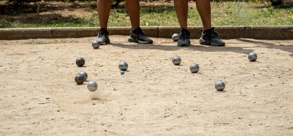 Metal ball from the game of petanque approaching the bowling alley bouncing off the sandy ground raising dust from a petanque court on a sunny day with opposing players in the background
