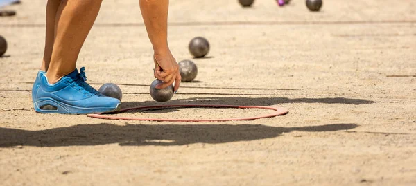 Spor ayakkabılı genç bir kadının eli, kumlu bir Petanque kortunda, güneşli bir günde Petanque oyunu oynamak için metal petanque topunu yakalıyor.