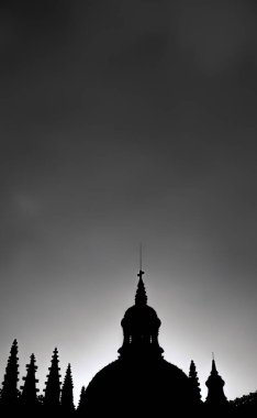Chiaroscuro and silhouette of Segovia Cathedral at sunset backlit by the sun with pigeons on rooftops and an ominous dramatic gray stormy sky in vertical format vision