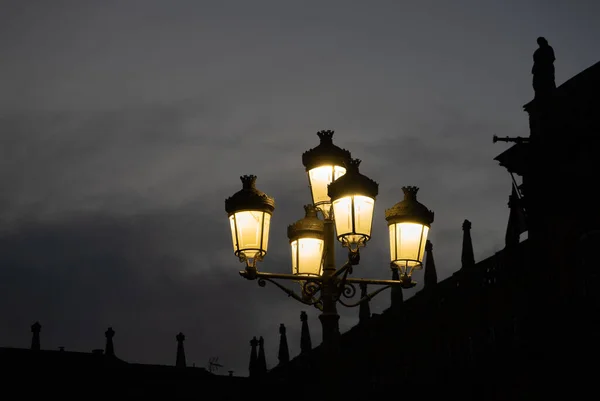 Wrought iron lamp with five lanterns dimly illuminating with yellow light at dusk in the main square of Salamanca with the gray and dramatic sky and the silhouette of the palace and sculptures