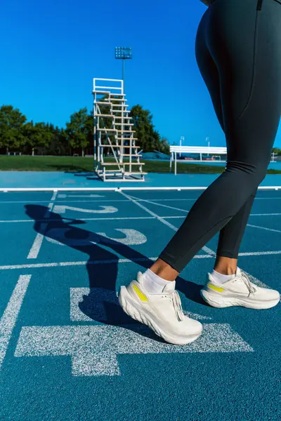 Shadow cast on a blue running track of a tanned young female runner, prepared to do a sprint running with her legs bent and in position on a blue track.