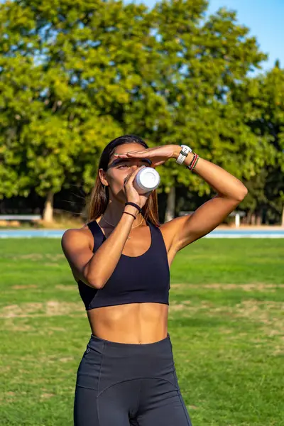 Beautiful young runner, tanned with long dark hair, protecting her eyes from the sun with her hand, looking at the horizon and drinking and hydrating herself with a can of isotonic drink.