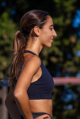 Güzel ve narin bir kız, sıkı spor kıyafetleri içinde, elleri belinde ve sakince gülümseyen ve mutlu bir şekilde maraton yarışından önce bekliyor..