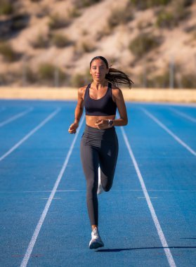 Güzel, ince, bronzlaşmış kadın koşucu, sıkı spor kıyafetleri içinde, mavi bir pistte koşmaktan zevk alıyor, kameraya yaklaşıyor ve bakıyor..