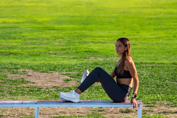 Dar spor kıyafetleri giymiş, uzun saçlı, ahşap bir bankta oturan, koşan bir yarıştan önce dikkatle ufka bakan güzel bir genç kız..