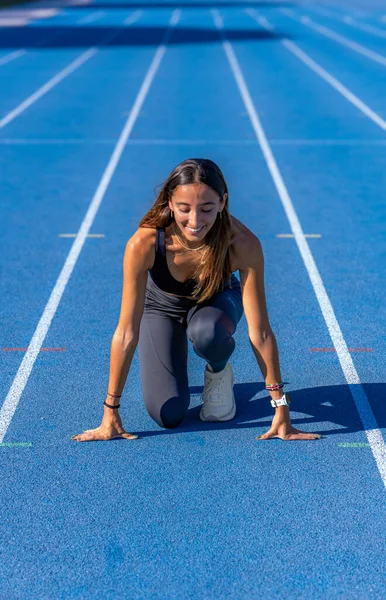 Beautiful young runner girl, tanned with long dark hair, smiling looking at the track at the starting line of a blue running track with her hands resting on the ground and legs bent to run.