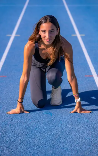Beautiful young runner girl, tanned with long dark hair, smiling looking at camera at the starting line of a blue running track with her hands resting on the ground and legs bent to run.