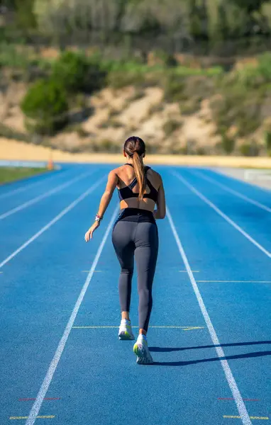 Güzel, bronzlaşmış, genç, sımsıkı spor kıyafetleri giymiş, bacakları bükülmüş ve mavi bir atletizm pistinde koşmaya hazır bir kız..