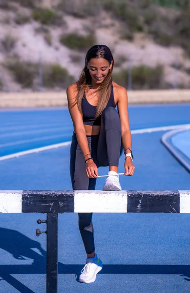 Beautiful young runner girl, tanned with long brown hair, smiling with her leg bent leaning on an obstacle on a running track tying the laces of her sports shoes.
