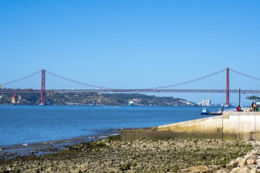 Lizbon 'un kırmızı metal 25 de Abril çelik asma köprüsünün panoramik manzarası. Plaj sahilinden Golden Gate gibi. İnsanlar şemsiyeli bir barda oturuyor. Limanda mahsur kalmış bir turist teknesi.