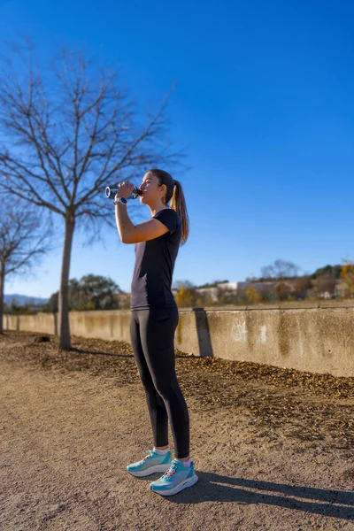 Beautiful young runner girl, beautifully illuminated by sunrise light, drinking water and hydrating from a bottle, dressed in tight black sportswear, looking at the horizon in a park.