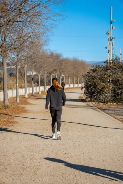 Young woman with a ponytail, dressed in sports clothing and black tights, walking quickly along a path in a public park to consume calories and burn fat. People walking.