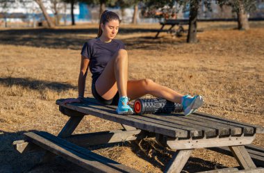 Genç sporcu kız, müzik dinliyor ve tahta bir masaya yaslanıyor. Baldırının arkasında, engebeli siyah köpüklü vücut masajı silindiri ile masaj yapıyor..