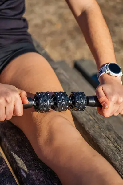 Young sportsman in running shorts, practicing self-massage on the quadriceps part of his leg with a bumpy black foam body massage roller, on a wooden bench in a mountain park in the morning.